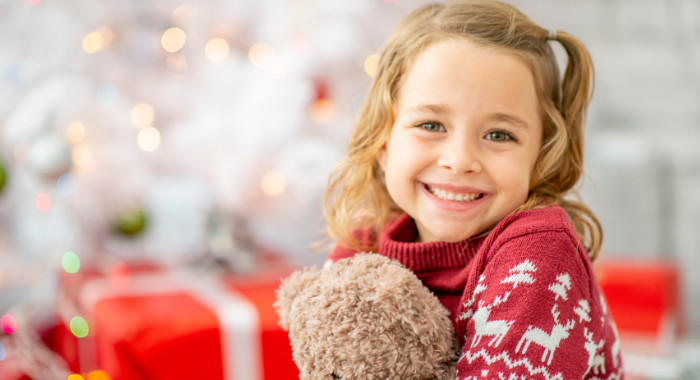 Little girl sitting in front of holiday decorations with presents and holding a bear.