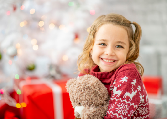 Little girl sitting in front of holiday decorations with presents and holding a bear.