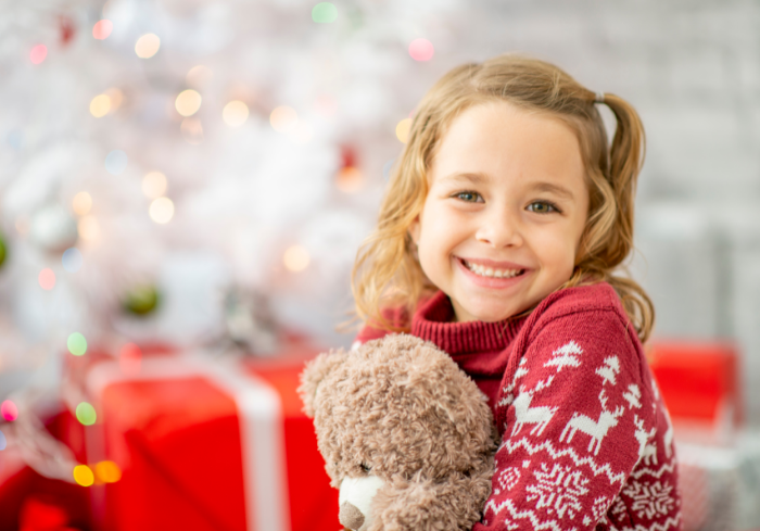 Little girl sitting in front of holiday decorations with presents and holding a bear.